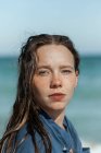 Donna in camicia bagnata e con i capelli bagnati in piedi guardando la fotocamera sulla spiaggia vicino al mare mentre si gode la giornata estiva — Foto stock