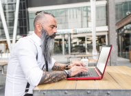 Vista lateral do trabalhador masculino hipster pensativo em netbook de navegação de desgaste formal enquanto sentado à mesa com café no terraço da cafetaria — Fotografia de Stock