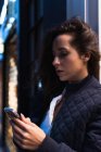 Vista lateral de la mujer reflexiva en ropa de abrigo utilizando el teléfono inteligente mientras está de pie en la calle y mirando a la pantalla por la noche - foto de stock