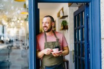 Barista mâle barbu positif dans le tablier avec tasse de café chaud dans les mains debout dans l'embrasure de la porte du café moderne — Photo de stock