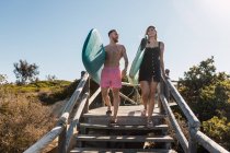 Corpo completo di coppia sportiva con tavole da surf passeggiando sulle scale insieme su un sentiero di legno vicino a piante verdi prima dell'allenamento in località tropicale — Foto stock