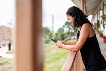 Side view of cheerful young ethnic lady with curly dark hair in casual clothes smiling while messaging on mobile phone standing on wooden terrace of aged house in countryside — Stock Photo