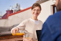 Вид сбоку на положительную молодую женщину в очках, наливающую свежий сок из стеклянной бутылки в чашку соседа-мужчины, работающего на ноутбуке на балконе — стоковое фото