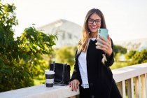 Позитивна жінка в стильному формальному вбранні, що стоїть з ноутбуком і бере самопортрет на мобільний телефон біля перила моста — стокове фото