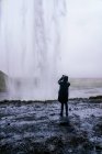 Back view of unrecognizable traveler in warm outerwear and hoodie taking photo of picturesque Seljalandsfoss waterfall on smartphone during trip in Iceland — Stock Photo