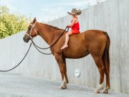 Visão lateral corpo inteiro do menino descalço em chapéu sentado no cavalo castanho e segurando rédea — Fotografia de Stock