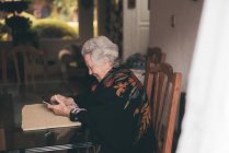 Vista lateral de una anciana sonriente vestida con ropa de abrigo sentada a la mesa con una tableta y una taza de té mirando la pantalla - foto de stock