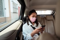 Passageira étnica com cinto de segurança apertado usando o telefone celular enquanto montava máscara protetora no banco de trás no táxi — Fotografia de Stock