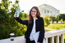 Позитивна жінка в стильному формальному вбранні, що стоїть з ноутбуком і бере самопортрет на мобільний телефон біля перила моста — стокове фото