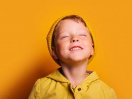 Счастливый мальчик в ярко-желтой куртке и шляпе-шапочке смеется с закрытыми глазами на жёлтом фоне в студии — стоковое фото
