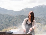 Concentrated female chef with tongs in apron cooking sausages on grill grate while standing on grassy field in countryside — Stock Photo
