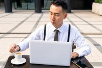 Wistful jovem empresário asiático com xícara de bebida quente e netbook na mesa de cafetaria urbana à luz do dia — Fotografia de Stock