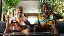 From below cheerful multiracial friends with gamepads in hands sitting on couch while playing video game together in light living room with green plant — Stock Photo