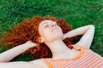 Top view of happy curly haired female with hands behind head relaxing on lawn with eyes closed — Stock Photo