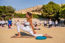 Side view of calm woman in activewear performing crescent lunge on knee with hands interlaced on mat during yoga practice with instructor in park in sunny day — Stock Photo