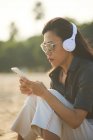 Seitenansicht einer ernsthaften Asiatin mit Sonnenbrille, die SMS auf dem Handy sendet, während sie am Sandstrand über Kopfhörer Musik hört — Stockfoto