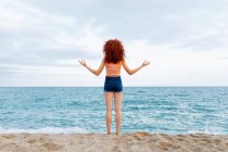 Vue arrière d'une femelle méconnaissable aux cheveux roux bouclés faisant un geste zen sur le rivage d'une mer bleu ondulant — Photo de stock