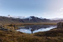 Visão traseira de turista masculino irreconhecível em roupas casuais admirando a natureza selvagem enquanto está perto de lago pacífico refletindo montanhas nevadas na Islândia — Fotografia de Stock