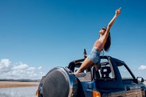 Full body of happy young female in summer outfit and sunglasses raising hand of beer while standing on roof of safari car on shore of river — Stock Photo