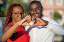 Щаслива афроамериканська пара, яка обіймає і показує знак у формі серця руками, стоячи на вулиці в сонячний день. — стокове фото