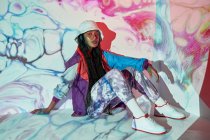 Сверху молодая доминиканская девушка-подросток в модном наряде и шляпе сидит возле белой стены с творческими абстрактными проекциями и смотрит в камеру — стоковое фото