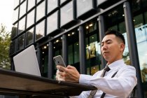 Знизу молодий етнічний чоловік-підприємець з кавою серфінг Інтернет на мобільному телефоні, сидячи за міським столом кафетерії з ноутбуком — стокове фото