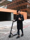 Giovane maschio afroamericano elegante con capelli ricci scuri in elegante cappotto in piedi sulla strada con scooter elettrico e tazza di caffè da asporto e guardando altrove — Foto stock