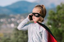 Menina auto-assegurada no traje de super-herói máscara olho com capa ter telefonema no celular — Fotografia de Stock