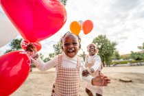Irmãzinhas afro-americanas felizes em vestidos semelhantes em pé com balões coloridos nas mãos na grama verde no parque à luz do dia — Fotografia de Stock