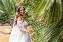 Позитивная женщина с вьющимися волосами в белом свадебном платье смотрит в сторону, стоя рядом с экзотическими зелеными деревьями во время празднования праздника — стоковое фото