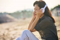Vista lateral de pacífica mujer asiática con los ojos cerrados escuchando la canción de los auriculares inalámbricos mientras se sienta en la orilla arenosa - foto de stock