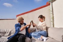 Entzückte junge Freunde in lässiger Kleidung sitzen auf dem Sofa und klirren an Bierflaschen, während sie gemeinsam auf der Terrasse chillen — Stockfoto