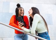Снизу веселые разнообразные женщины в стильной одежде, стоящие на лестнице возле перила и смеющиеся во время просмотра смартфона на улице в дневное время — стоковое фото