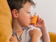 Side view of little boy with asthma using inhaler while sitting on sofa at home — Stock Photo