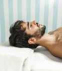 Vista laterale del maschio barbuto senza maglietta sdraiato sul lettino da massaggio con gli occhi chiusi — Foto stock
