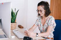 Glad femelle en tenue décontractée et lunettes assis au bureau avec ordinateur et smartphone de navigation — Photo de stock
