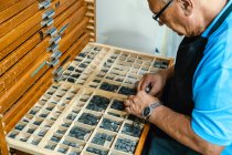 Зверху врожаю зосереджений старший чоловічий майстер в фартухах і окулярах вибираючи друк прес-літер з дерев'яної коробки під час роботи в традиційному ательє — стокове фото