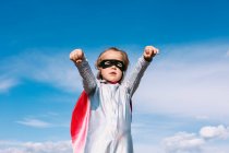 Знизу маленька дівчинка в костюмі супергероя піднімає витягнуті кулаки для показу влади, стоячи на тлі блакитного чистого неба — стокове фото
