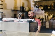 Durch das Glas eines männlichen Arbeiters in der Schürze, der während der Arbeit in der modernen Cafeteria am Fenster steht und Kaffee zubereitet — Stockfoto
