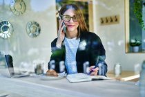 Через стекло деловая женщина разговаривает на смартфоне за столом с кофе с круассаном возле ноутбука во время работы удаленно — стоковое фото