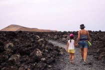Back view of anonymous mom with girl holding hands while strolling on pathway between lava against Caldereta and Caldera Blanca Volcanoes in Spain — Stock Photo
