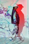 Seitenansicht einer selbstbewussten jungen dominikanischen Teenagerin in trendigem Outfit und Hut, die in der Nähe einer weißen Wand mit kreativen abstrakten Projektionen steht und in die Kamera blickt — Stockfoto