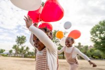 Щасливі афроамериканські маленькі сестри в схожих сукнях стоять з барвистими кульками в руках на зеленій траві в парку вдень. — стокове фото