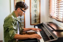 Vista laterale del musicista maschile in abiti casual che suona la melodia al pianoforte mentre è seduto in un appartamento moderno vicino alla finestra — Foto stock
