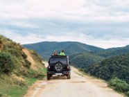 Back view of unrecognizable travelers standing in SUV car driving along rough road during trip in picturesque mountainous valley — Stock Photo