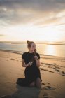 Serious female with black feather wearing dress kneeling on sandy dune washed by sea at sundown — Stock Photo