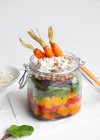 Салат з різнокольоровим стиглим подрібненим болгарським перцем і булгуром увінчаний сирою морквою, поданою в банці на столі на білому тлі — стокове фото