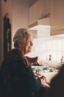 Seitenansicht einer glücklichen positiven Seniorin mit grauen Haaren, die warme Kleidung trägt und in der Küche an der Spüle steht — Stockfoto