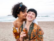 Loving multiethnic couple of females drinking beer and enjoying summer day on seashore — Stock Photo