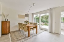 Modernes Esszimmerinterieur mit Stühlen und Holztisch auf Teppich unter Lampe gegen Fensterwand zu Hause — Stockfoto